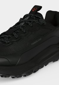 Μαύρο αθλητικό παπούτσι Timberland με υφασμάτινη υφή, παχιά σόλα, κορδόνια με σχέδια και ραβδί για κράτημα με πορτοκαλί λογότυπο σε λευκό φόντο.