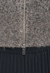 Κοντινό πλάνο από μια ασημένια φερμουάρ κλεισμένη ανάμεσα σε γκρι πλεκτό ύφασμα και μαύρο πλισέ υλικό.