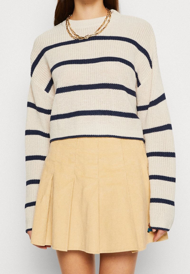 Femme portant un pull crème avec des rayures horizontales bleu marine, un collier chaîne doré et une jupe plissée beige.