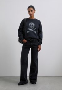 Sudadera negra con gráfico de calavera y tibias cruzadas, combinada con pantalones vaqueros de pierna ancha negros. Modelo sosteniendo un bolso negro. Textura suave, diseño casual.