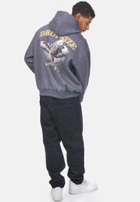 Sudadera gris con un gráfico de un águila y texto en la parte posterior. Combinada con pantalones negros y zapatillas deportivas blancas. Presenta una capucha con cordón y puños acanalados.