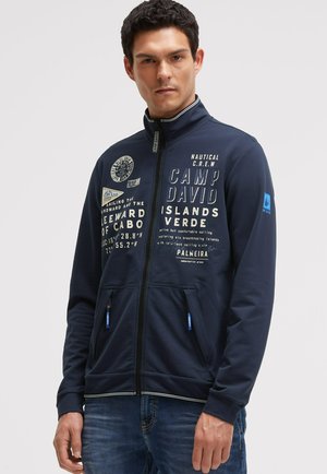 Man wearing navy blue zip-up jacket with white nautical-themed text and patches, standing against light grey background.