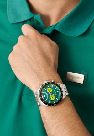 Orologio da polso argentato con quadrante verde, dotato di due sottocadrani gialli e dettagli neri. Indossato su una mano che tiene una maglietta polo verde.