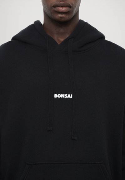 Črna kapuca s kenguru žepom, vrvico in belim logotipom "BONSAI" na prsih. Narejena iz mehkega materiala z gladko teksturo.