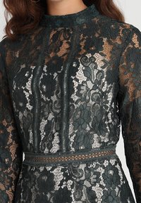 Robe en dentelle avec un col haut et des manches longues, de couleur vert foncé, ornée de motifs floraux et de détails texturés le long de la taille.