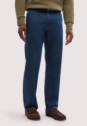 Mand iført mørkeblå jeans med lige ben, brune ruskindssko og en olivengrøn sweatshirt, stående mod en lys baggrund.