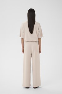 Hosszú sötét hajjal rendelkező nő, laza, bézs színű, rövid ujjú felsőt és bő szárú nadrágot visel, háttal áll egy fehér háttér előtt.