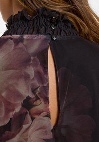 Zwarte mesh top met een gerimpelde halslijn en een opening aan de achterkant, vastgemaakt met drie zwarte knopen, met bloemenprint details.