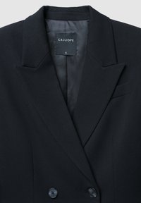 Blazer nero doppiopetto realizzato in tessuto liscio, con rever a punta, due bottoni frontali e una piccola tasca sul petto. Etichetta del marchio visibile.