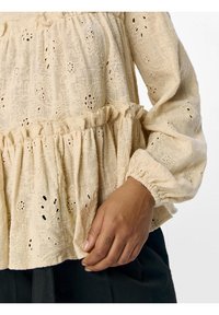 Béžová vyšívaná blúzka s peplum strihom, dlhými rukávmi, naťahovanými manžetami a zložitými kvetinovými vzormi. Tkanina s textúrou a detaily v tvare očiek.