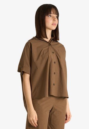 Giovane donna con capelli castani lisci, indossa una camicia marrone a maniche corte e ampie con bottoni e pantaloni abbinati, in piedi davanti a uno sfondo bianco.