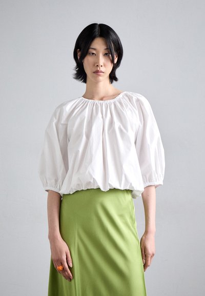 Blouse blanche à manches bouffantes avec un col froncé et jupe verte. Texture de tissu douce avec une finition brillante. Accessoire en anneau orange à la main.