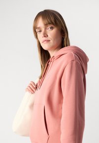 Felpa con cappuccio in morbido cotone rosa chiaro, dotata di tasca frontale e cordini regolabili. Il modello tiene una borsa a tracolla color crema.
