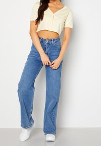 Krótka, jasnżółta koszula na guziki z krótkimi rękawami, zestawiona z dżinsami o wysokim stanie i szerokich nogawkach. Całość uzupełniają białe sneakersy.