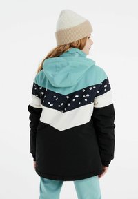 Veste colorée en blocs de teal, noir et blanc avec un motif floral sur fond marine. Elle possède une capuche et des poignets élastiques. Portée avec un bonnet tricoté crème.