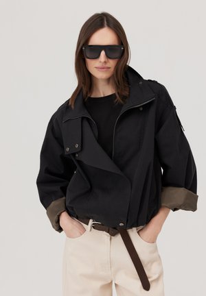 Kobieta nosząca czarną, oversize’ową kurtkę ze związanymi rękawami, ciemne okulary przeciwsłoneczne, beżowe spodnie i brązowy pasek, ręce w kieszeniach.
