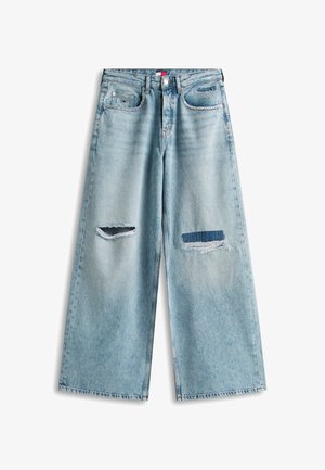 Φαρδιά τζιν σε ανοιχτό μπλε χρώμα με σκισίματα, συμπεριλαμβανομένης μίας σχισμής στο αριστερό μηρό και ενός μπαλώματος σε δάκρυ στο δεξί μηρό.