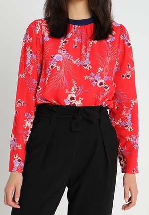 Blusa floreale rossa con maniche lunghe, scollo arricciato e bordini scuri, abbinata a pantaloni neri a vita alta con dettaglio della cintura.