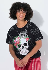 T-shirt graphique noir avec un grand crâne entouré de roses roses et d'une couronne, présentant une texture usée. Associé à un pantalon rayé rouge.