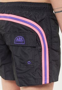 Pantaloni da bagno neri con cintura in vita elastica, caratterizzati da strisce laterali rosa e viola e un patch viola SUNDEK su una tasca posteriore.