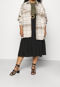 Rutet knap-up skjorte i beige og brun, olivengrøn top, sort lagdelt nederdel, og sorte sandaler med blokhæl og stropper.