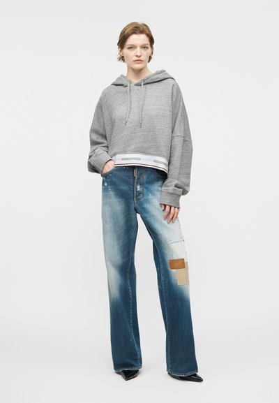 Νεαρό άτομο που φοράει γκρι κοντό φούτερ με κουκούλα και φαρδιά μπλε ξεθωριασμένα τζιν με μπαλώματα, στέκεται με το ένα χέρι στην τσέπη μπροστά σε λευκό φόντο.