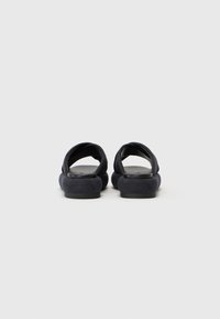 Mørkeblå sandaler med glatt tekstur, åpen bak, og rund tå-design. Polstrede stropper krysser over toppen for komfort. Flatt såle.