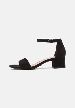 Sandale noire en daim à talon bas carré, bout ouvert, bride à la cheville et boucle latérale sur fond blanc uni.
