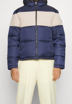 Persona che indossa una giacca imbottita blu navy e beige con cerniera frontale e pantaloni color crema, in piedi contro uno sfondo semplice.