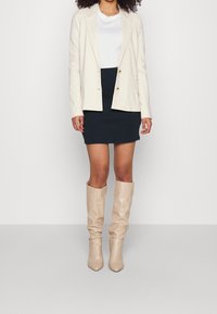 Crème blazer met gouden knopen, gecombineerd met een marineblauwe rok en beige kniehoge laarzen. Eenvoudig ontwerp, gladde textuur en getailleerd silhouette.