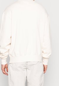 Felpa off-white con colletto a girocollo, spalle scese e polsini a costine. Caratterizzata da un tessuto morbido e testurizzato, ha una vestibilità rilassata.