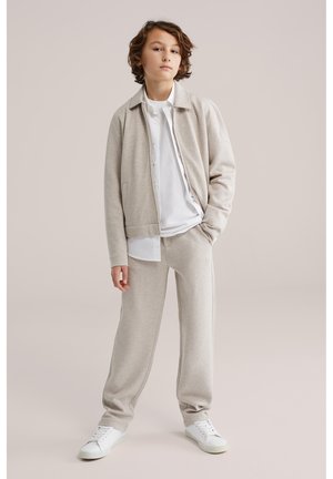 Fiatal személy áll egy kézzel a zsebében, bézs dzsekit és nadrágot, fehér inget és fehér tornacipőt viselve egyszínű háttér előtt.