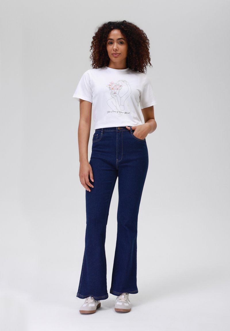 T-shirt bianco cropped con design floreale in linea, abbinato a jeans a vita alta blu scuro a palazzo e sneakers beige, in piedi su uno sfondo neutro.
