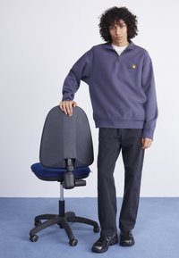 Lila fleece-tröja med kvartdragkedja och broderad logga, ihop med svarta jeans och svarta sneakers, stående vid en kontorsstol.