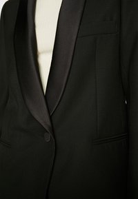 Veste de smoking noire avec un revers en satin, des épaules structurées et une fermeture à un seul bouton. Texture lisse avec une coupe ajustée.
