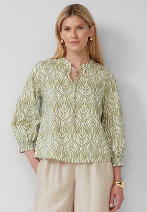 Blouse vert clair avec un motif de feuilles blanches, décolleté rond et détail à nouer. Manches longues avec poignets élastiques. Associée à un pantalon couleur crème.