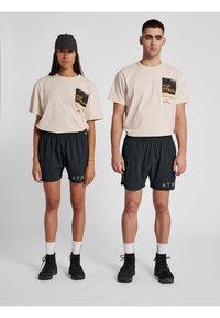 Beżowe, oversize'owe t-shirty z graficznym wzorem kieszeni, sparowane z czarnymi szortami z elastycznym paskiem i logo "ATW". Czarne obuwie.