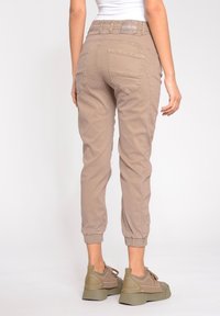 Pantalones jogger beige con puños elásticos en los tobillos, bolsillos traseros y un parche con logo en la cintura. Llevados con zapatillas de color verde olivo.