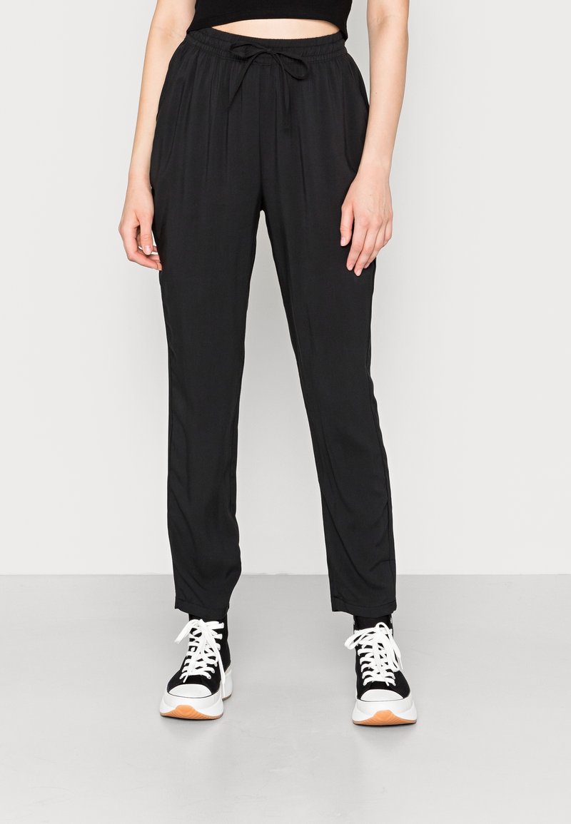 Personne portant un pantalon noir à cordon, un crop top noir et des baskets noires et blanches, debout devant un fond clair uni.