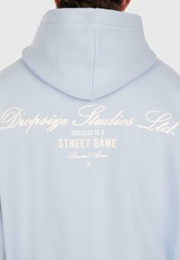 Person, der einen hellblauen Hoodie trägt, auf dem hinten der Text "Dropsize Studios Ltd. Erfolg ist ein Straßenspiel mit eingeschränktem Zugang" gedruckt ist.