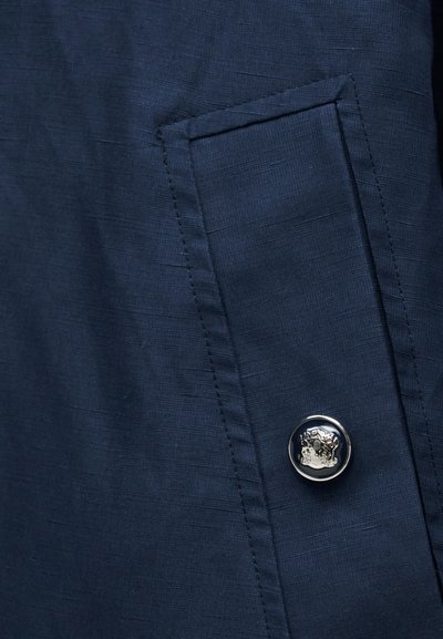 Σκούρο μπλε ύφασμα με ορθογώνια τσέπη με καπάκι, ασφαλισμένη με ένα μόνο στρογγυλό μεταλλικό κουμπί χαραγμένο με σχέδιο θυρεού.