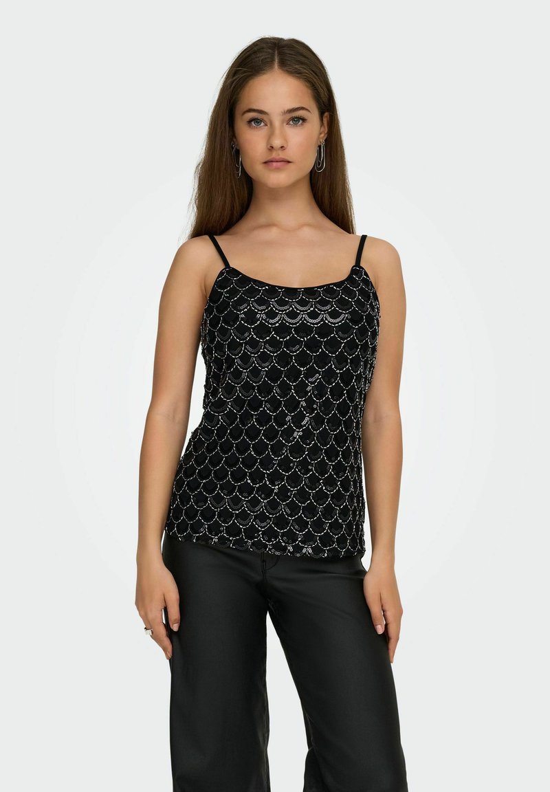 Jeune femme aux longs cheveux portant un haut sans manches noir à motif pailleté et un pantalon noir, debout devant un fond clair uni.