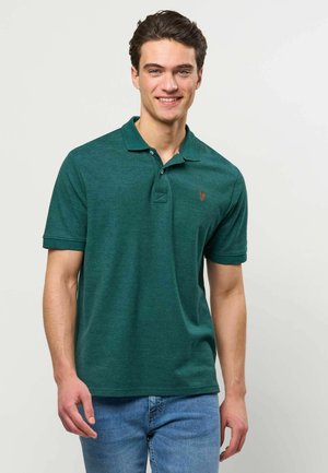 Jeune homme souriant portant un polo vert à manches courtes et un jean bleu, debout devant un fond clair uni.