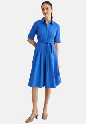 Mujer vestida con un vestido azul de manga corta, con cuello, hasta la rodilla, con cinturón anudado en la cintura y zapatos beige de tacón ancho, de pie sobre un fondo blanco.
