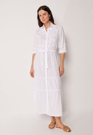 Frau trägt ein weißes, langärmeliges Midikleid mit Blumenstickerei und Taillengürtel, kombiniert mit beigefarbenen Slide-Sandalen.