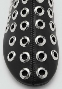 Zapato de cuero negro con punta redondeada, que presenta ojales plateados distribuidos uniformemente por toda la superficie y un detalle de costura central.