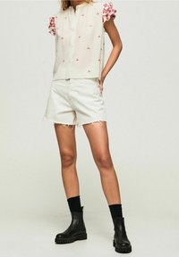 Donna che indossa una camicia bianca con bottoni e ricami rossi, pantaloni corto bianchi sfilacciati, calze nere e stivali neri con suola spessa, in piedi con una mano in tasca.