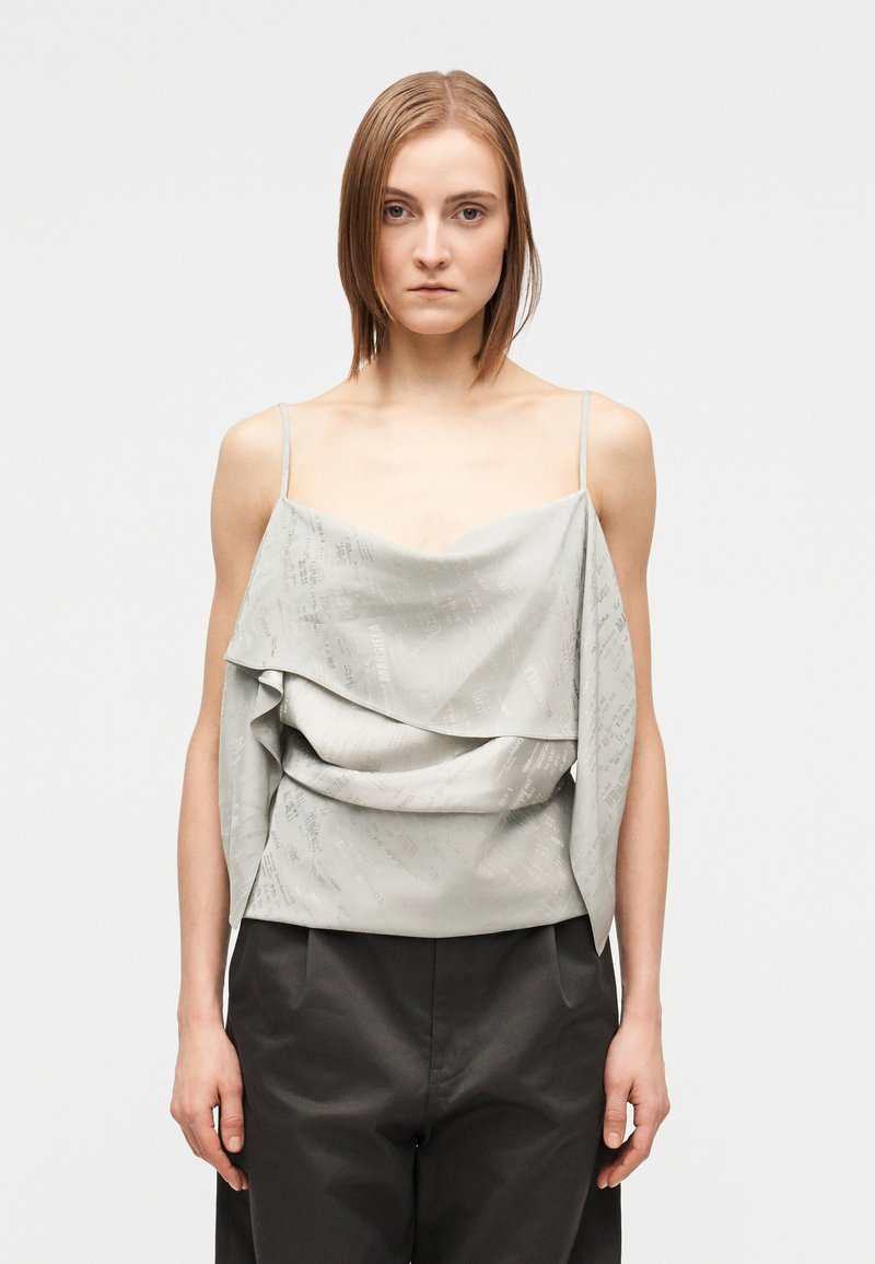 Ženska s ravnimi lasmi nosi svetlo siv brezrokavnik z nagubanim materialom in črne hlače z visokim pasom, stoji pred belo enobarvno podlago.