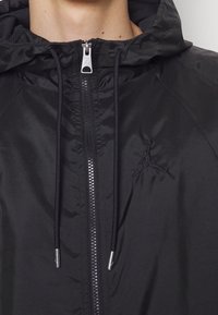 Svart huva jacka i slätt, lätt material med dragkedja fram, utrustad med en broderad logotyp och snoddar vid huvan.