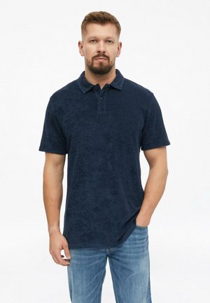 Man met kort haar en baard, gekleed in een donkerblauw poloshirt met korte mouwen en lichtblauwe jeans, staand met één hand in de zak.